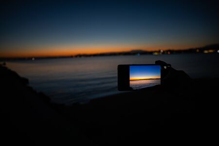 human hand holding a phone taking a picture of a sunset view over the seaの写真素材
