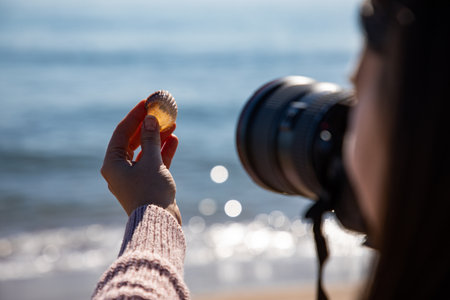Beautiful woman taking a picture of a sea shell holding it with her hand close back shot at the beachの写真素材