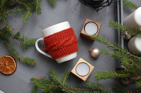 White and red ceramic mug on gray textile with candles and spruce branches