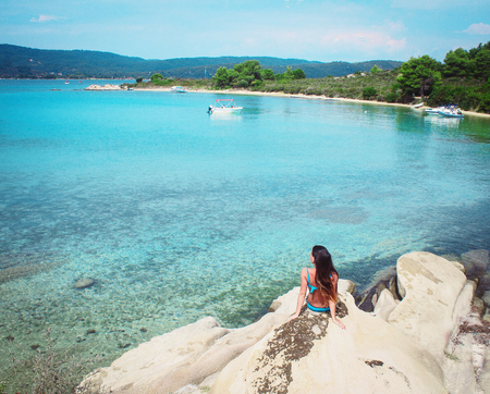 young brunette caucasian girl in blue swimsuit sitting on rocks and looking at turquoise blue waters of Aegean sea on sunny summer day, Chalkidiki, Greeceの写真素材