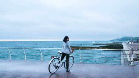 Woman in sport clothing riding white vintage bicycle on seaside during cloudy rainy day. Lady standing under water, wave or rain with emotionsの写真素材