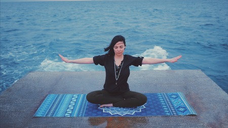 Meditation. Yoga young calm brunette woman by the waves sea on pier relaxing in serene zen. lotus yoga pose on a beach at cloudy weather. Beautiful colorful background.の写真素材