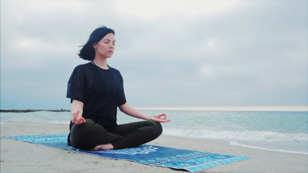 Fitness woman in sports clothing practicing yoga on the sea ocean beach close up to water. Dolly shot. Calm young woman sitting in lotus pose.の写真素材