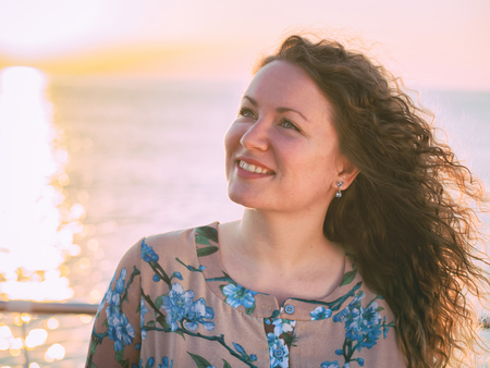 Portrait of young beautiful woman with curly blond hair standing near sea or ocean in sunriseの写真素材