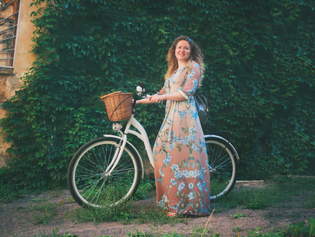 Beautiful young attractive woman standing near white retro bicycle on green wall with leaves background.の写真素材