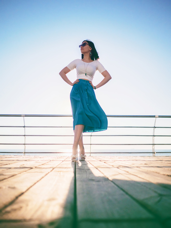 Beautiful young woman posing as model standing on embankment near sea or ocean. Lady wearing retro style skirt and sunglasses. Morning sunlight.の写真素材
