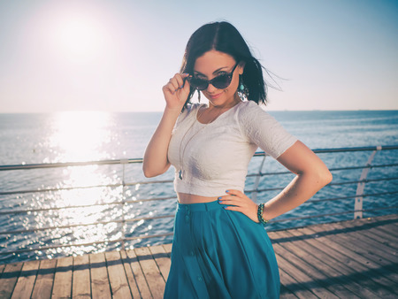 Beautiful young woman posing as model standing on embankment near sea or ocean. Lady wearing retro style skirt and sunglasses. Morning sunlight.の写真素材