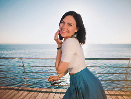 Beautiful young woman posing as model standing on embankment near sea or ocean. Lady wearing retro style skirt and sunglasses. Morning sunlight.の写真素材