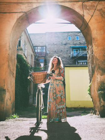 Beautiful young woman wearing long dress standing near retro white bicycle with basket and looking on arch in old courtyardの写真素材