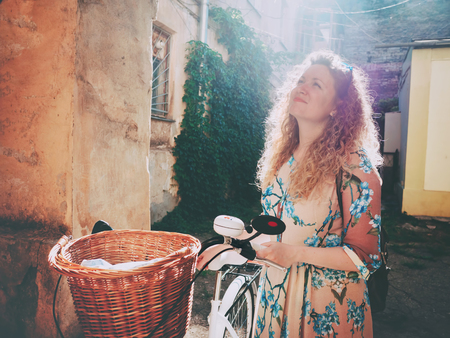 Beautiful young woman wearing long dress standing near retro white bicycle with basket and looking on arch in old courtyardの写真素材