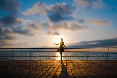 Silhouette of dancing ballerina in black ballet tutu and pointe on embankment above ocean or sea at sunrise or sunset. Young attractive blonde woman with long hair practicing stretching and exercises.の写真素材