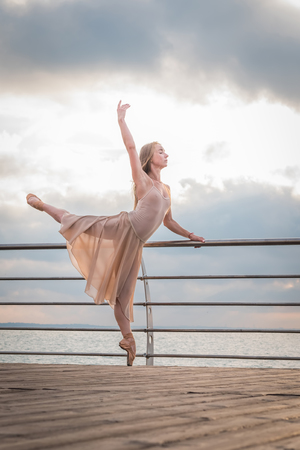 Dancing ballerina in beige silk dress and pointe on embankment above ocean or sea beach at sunrise. Young beautiful blonde woman with long hair practicing classic exercises with emotions.の写真素材