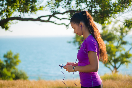 Female jogger listening music in headphones after training. Young athletic woman exercising in the park near to sea outdoors and enjoys music with earphones.の写真素材