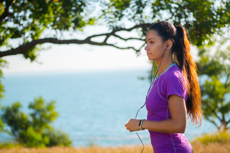 Female jogger listening music in headphones after training. Young athletic woman exercising in the park near to sea outdoors and enjoys music with earphones.の写真素材