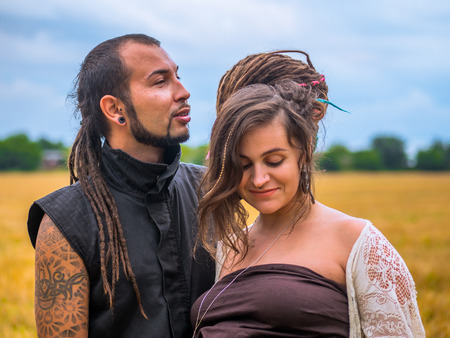 Man in a black cloth and pregnant woman in long dress with dreadlocks standing in wheat field on stormy sky background. Love story. Informal people with tattoo and piercing.の写真素材
