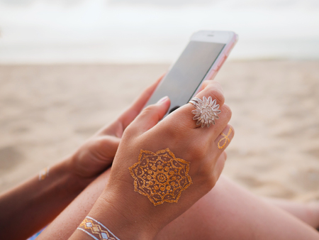 Woman with flash tattoo holding smartphone in hand on the beach. Boho style. Beautiful accessories.の写真素材