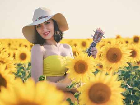 Happy woman in sunflower field playing on ukulele and singing. Summer girl in cowboy straw hat. Caucasian young lady have fun.. Little guitar. Music concept.の写真素材