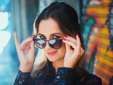 Close up portrait of pretty blonde young woman with glasses against colorful graffiti background. Female fashion model posing with sunglasses. Akcent on glassesの写真素材