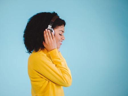 Beautiful young woman in headphones listening to music, smiling with closed eyes standing on a blue background in yellow sweatshirtの写真素材