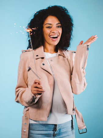 Sparkling Bengal fire in african american womans hand on blue background. Christmas Holiday Concept. Young pretty girl with afro hairstyle celebrating, smiling, enjoying timeの写真素材