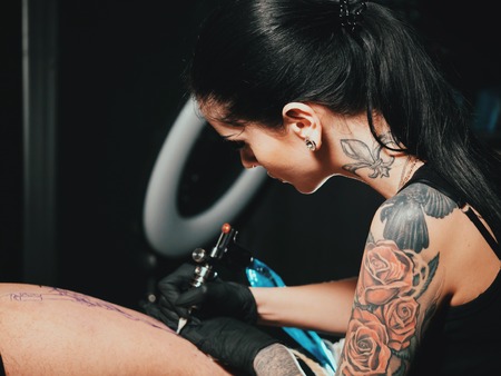 Close up tattoo artist demonstrates the process of getting black tattoo with paint. Concentrated female coloring contour with black ink on leg with gunの写真素材