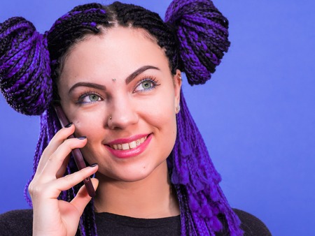 Adorable hipster caucasian woman with african colorful braids hairstyle talking with smartphone, using free wi-fi on mobile phone, smiling, posing on violet wall.の写真素材