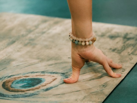 Beautiful young female hand with bracelets in sports clothes is stretching on yoga mat with peacock feature picture while working out at home or in studio. health concept. Focus on handsの写真素材