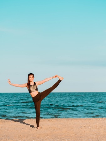 Young beautiful sporty woman in green clothing doing yoga asana on sea sandy beach near water. Girl practicing exercises. Health concept. Copy space.の写真素材