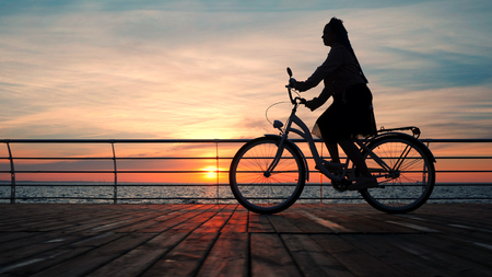 Wonderful sunrise or sunset above ocean. Silhouette of young stylish girl cycling on vintage bike on wooden embankment. Woman on bicycle near sea.の写真素材