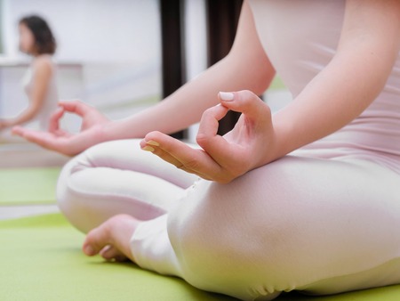 Young woman in beige jumpsuit sitting on green mat and practicing yoga at light studio class room. Meditating in lotus pose with Jnana Gian mudra. Om.の写真素材