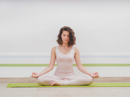Young pretty woman practicing in yoga at light minimalist studio class room. Attractive girl sits in classic lotus position and begins her meditationの写真素材