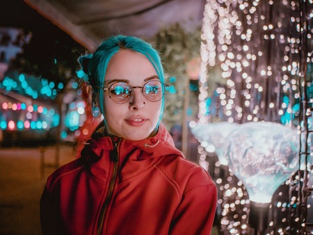 Young beautiful girl with blue dyed unusual hair, transparent glasses, piercing standing at night street with neon lights background. Portrait of happy cute stylish teenager.の写真素材