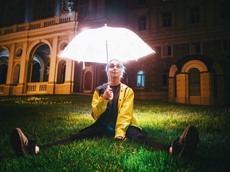 Young pretty girl with blue dyed hair in yellow raincoat and with transparent luminous umbrella...