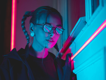 Young pretty girl with unusual hairstyle near glowing pink and blue neon lights of the city at night. Dyed blue hair in braids. Serious hipster teenager in glasses and beautiful lenses.の写真素材