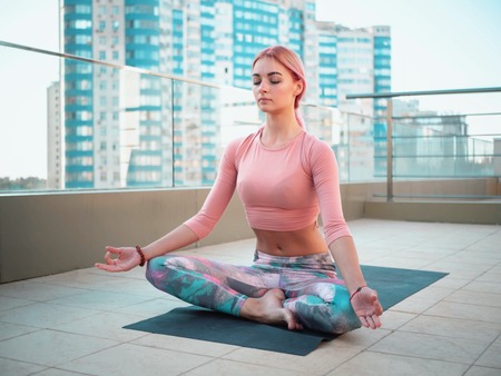 Young beautiful woman in pink tracksuit meditating during outdoor training on skyscraper terrace, she feeling peace in megalopolis. Sporty girl doing yoga on the roof.の写真素材