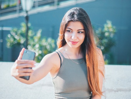 Portrait of young attractive female in fashion outfit making selfie outdoor while sitting on stairs. Beautiful woman with oriental face smiling to smartphone camera.の写真素材