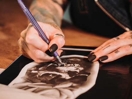 Tattoo artist drawing illustration of skull inside ink studio. Woman with black nails and rings at...