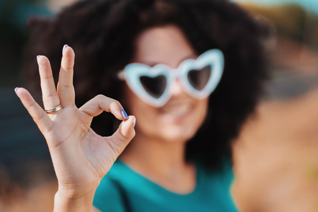Young pretty woman with african curly hairstyle and heart-shaped sunglasses showing OK sign. Beautiful girl outdoor.の写真素材