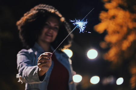 Young beautiful woman with very curly afro hair dancing with bengal fire at night illuminated street. Unusual trendy girl with sparklers. Holiday concept.の写真素材