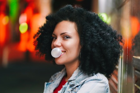 Young pretty woman with very curly afro hair inflates a bubble ball of white chewing gum at night. Trendy girl having fun, enjoying life. Beautiful illuminated street.の写真素材