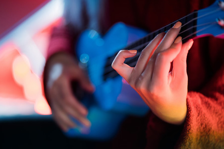 Hands of young pretty girl playing on blue ukulele while sitting on glowing neon stairs in amusement...