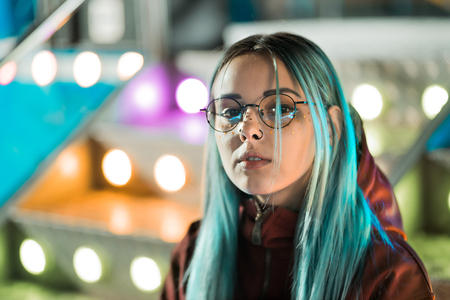 Hipster girl with blue dyed hair and golden sequins as freckles. Woman with nose piercing, transparent glasses, ears tunnels, unusual hairstyle stands in amusement night park.の写真素材
