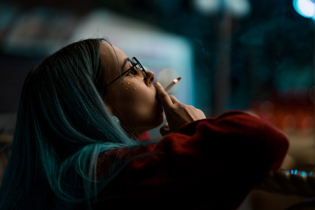Millennial cool pretty girl with unusual dyed hairstyle smoking cigarette near glowing neon wall at night. Blue hair, golden sequins as freckles,nose piercing. Mysterious hipster.の写真素材