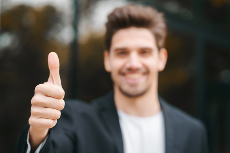 Winner. Success. Brunette young man in business clothing in office district smiles to camera and gives thumbs up. Happy guy showing gesture of approval.の写真素材