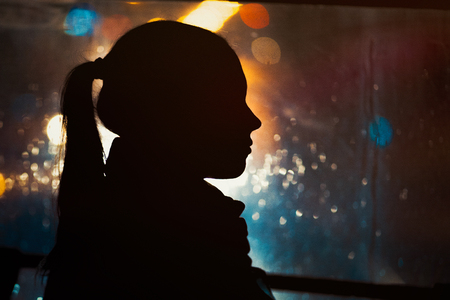 Silhouette of young girl on night city bokeh background. Rain drops running down the window. Autumn seasonの写真素材