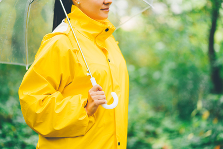 Attractive unrecognizable young girl in yellow raincoat in park or forest with transparent umbrella, autumn day. Focus on handsの写真素材