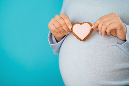 Pregnant woman holding heart-shaped cookie on tummy belly background. Young girl in blue wearing expecting baby. Maternity, motherhood, pregnancy, love concept. Copy spaceの写真素材
