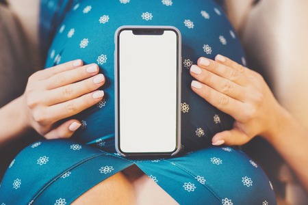 Unrecognizable pregnant woman holding her belly. Mobile phone with empty screen. Copy space