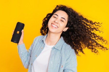 Modern trendy girl listening to music by wireless portable speaker. Young beautiful woman with curly...
