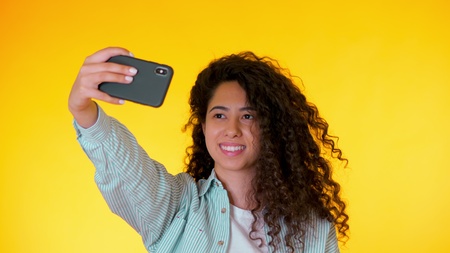 Beautiful woman with curly hair holding and using smart phone to film herself in yellow studio....